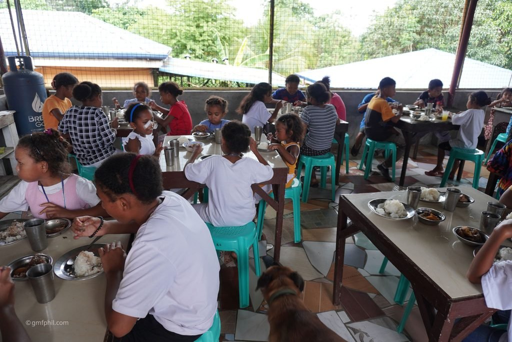 Many kids eating in lunch room. Long shot.